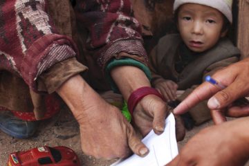 A small child looks at the camera while adults are being thumbprinted to vote in the census