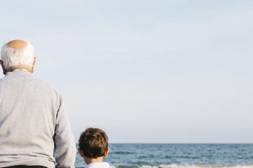 Back view of grandfather standing hand in hand on the beach with his grandchildren watching the sea