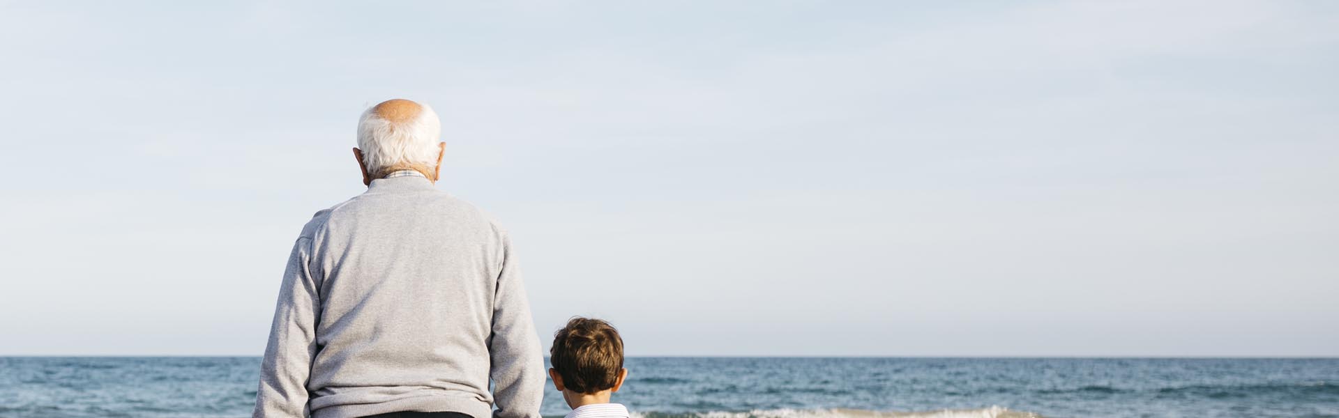 Back view of grandfather standing hand in hand on the beach with his grandchildren watching the sea