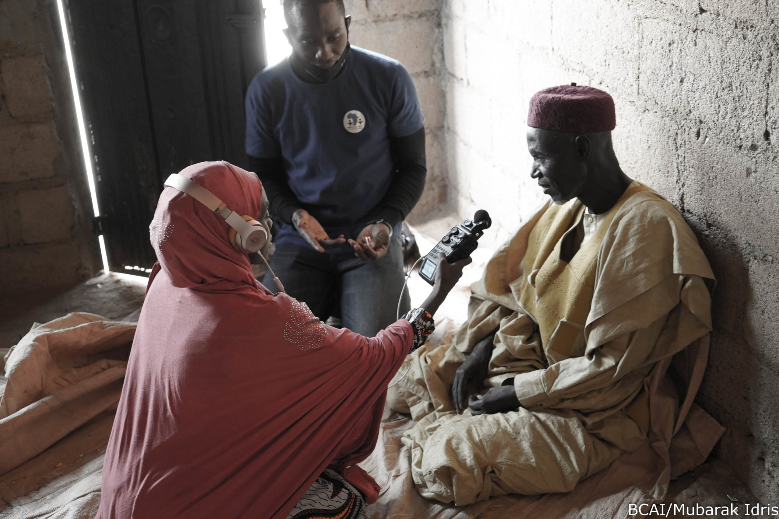 A muslim woman wearing headphones and holding a microphone interviews a man sitting on the ground in 