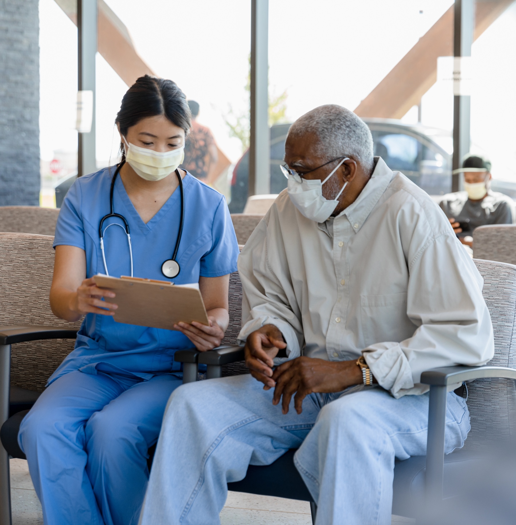 A nurse helps an elderly Black man wearing a mask fill out his paperwork in the lobby of a hospital.