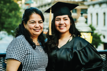A young Latina woman in a graduation cap and gown smiles with her mother.
