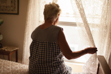 Elderly hispanic woman sits on her bed and looks out the window.