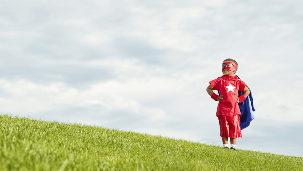 little boy in super hero costume with hands on hip looking at the sky smiling